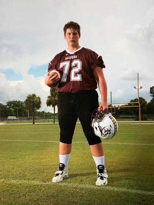 A teenage boy in a burgundy football uniform poses for a photo holding his helmet